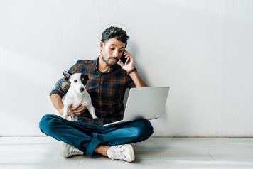 Younger man sitting on the floor with a dog in one hand while talking on the phone with the other and balancing a laptop on his lap.