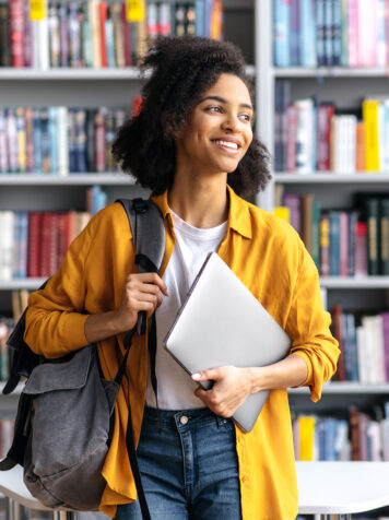 Portrait of joyful pretty african american female student standing against background of bookshelves in university library holding laptop and backpack looking to the side, smiling pleasant
